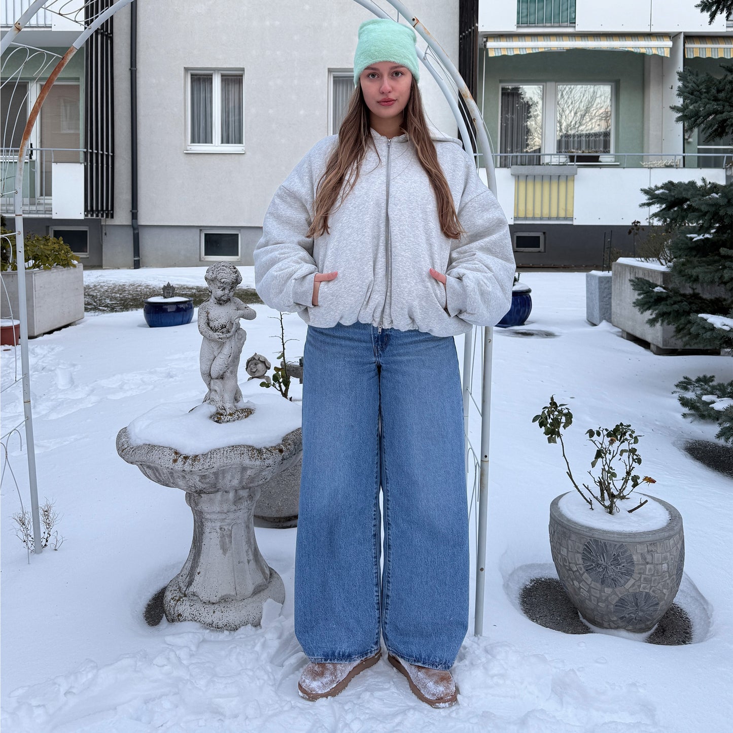 
                  
                    Full-length winter fashion shot of a women styled in a mint green fluffy beanie, oversized grey jacket, and blue wide-leg jeans in the snow.
                  
                