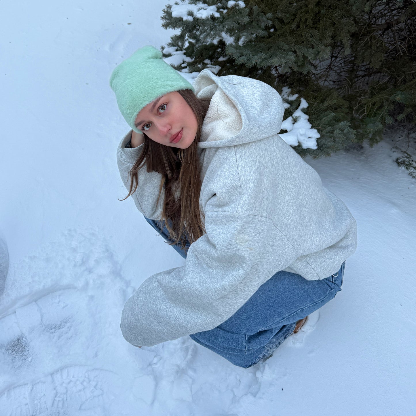 A Women wearing a cozy mint green fuzzy beanie and a grey hoodie in a snowy winter setting.